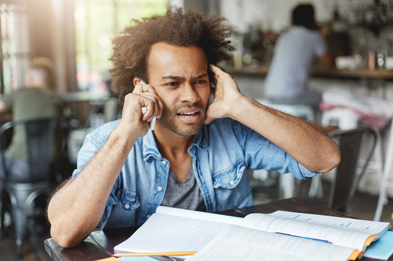 In a restaurant, a man has his hand over one ear while trying to hear the conversation on his phone.