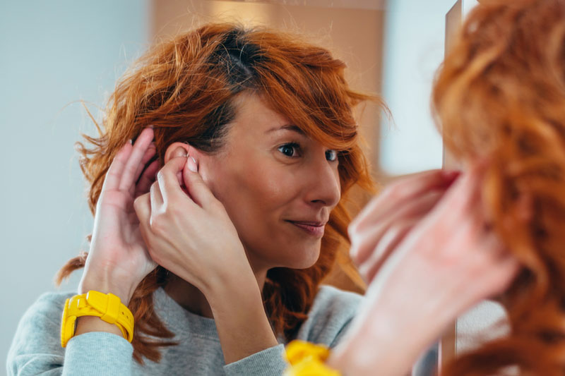 A redheaded woman is inserting a hearing aid into her ear and smiling while looking into a mirror.