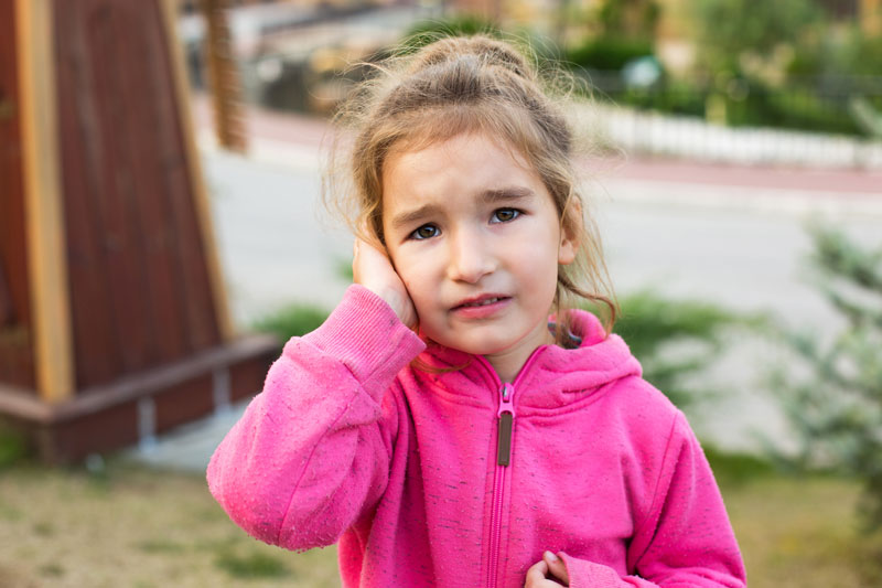 A small girl is standing outside, pouting with her hand covering her ear.