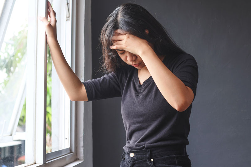A woman is at home and in pain, leaning against a wall with her hand over her forehead.