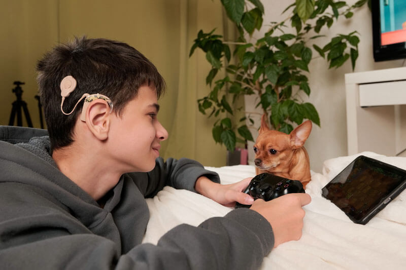 A teenage boy with a cochlear implant is laying down near a dog and playing video games.
