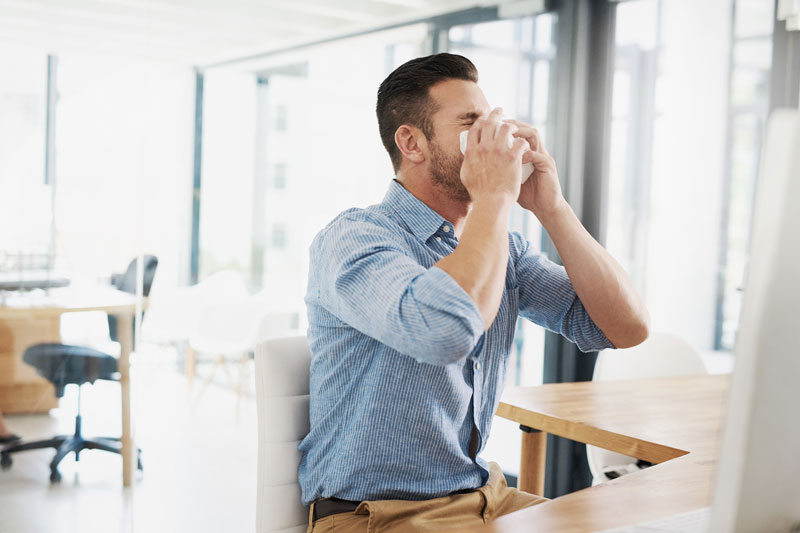 Young man at his desk, blowing his nose to relieve allergy pain