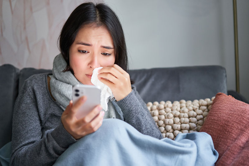 Young woman wipes nose with tissue while viewing her mobile phone in bed