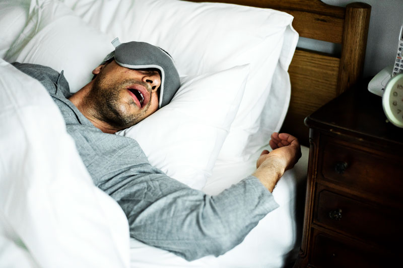 Man asleep on his back with his mouth open wearing an eye mask